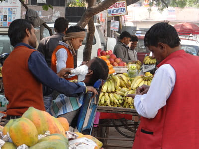 Inde, New Delhi : Dans la rue, un barbier rase un homme sur une chaise, devant un stand de vente de bananes. Des hommes sont autour.