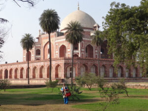 Inde, New Delhi : Un bâtiment en grès rouge avec des arches surmontées d'un dôme blanc et posé derrière des cocotiers et des arbres. Une femme pose devant pour la photo.