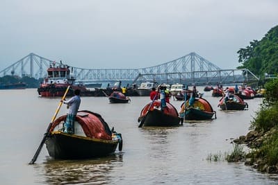 Inde : Des barques couvertes avec rameurs debout, sont sur un fleuve avec un pont en fer au loin