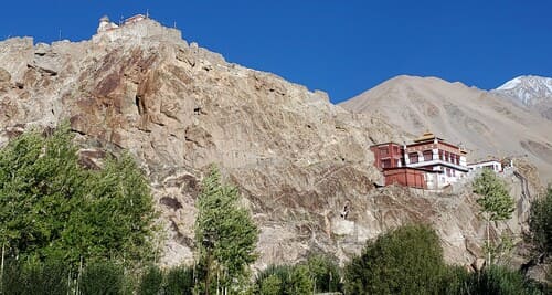 Ladakh : Un petit bâtiment rouge et blanc est accroché aux flancs d'une montagne. En bas de la photo, on aperçoit des arbres.